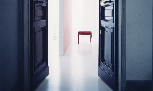 Vista dell’ingresso dal vano scale. Sul pavimento bianco spicca uno sgabello vintage con fodera rossa. Sulla mensola in alto: scatole di latta anni ‘60